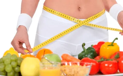 Uma mulher magra medindo seu abdomen com frutas na mesa, representando uma boa alimentação.