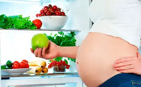 Uma gestante com a geladeira aberta segurando uma maçã, representando uma alimentação saudável.